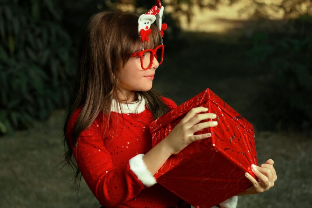 Young girl in red glasses and Santa headband holding a Christmas gift outdoors.